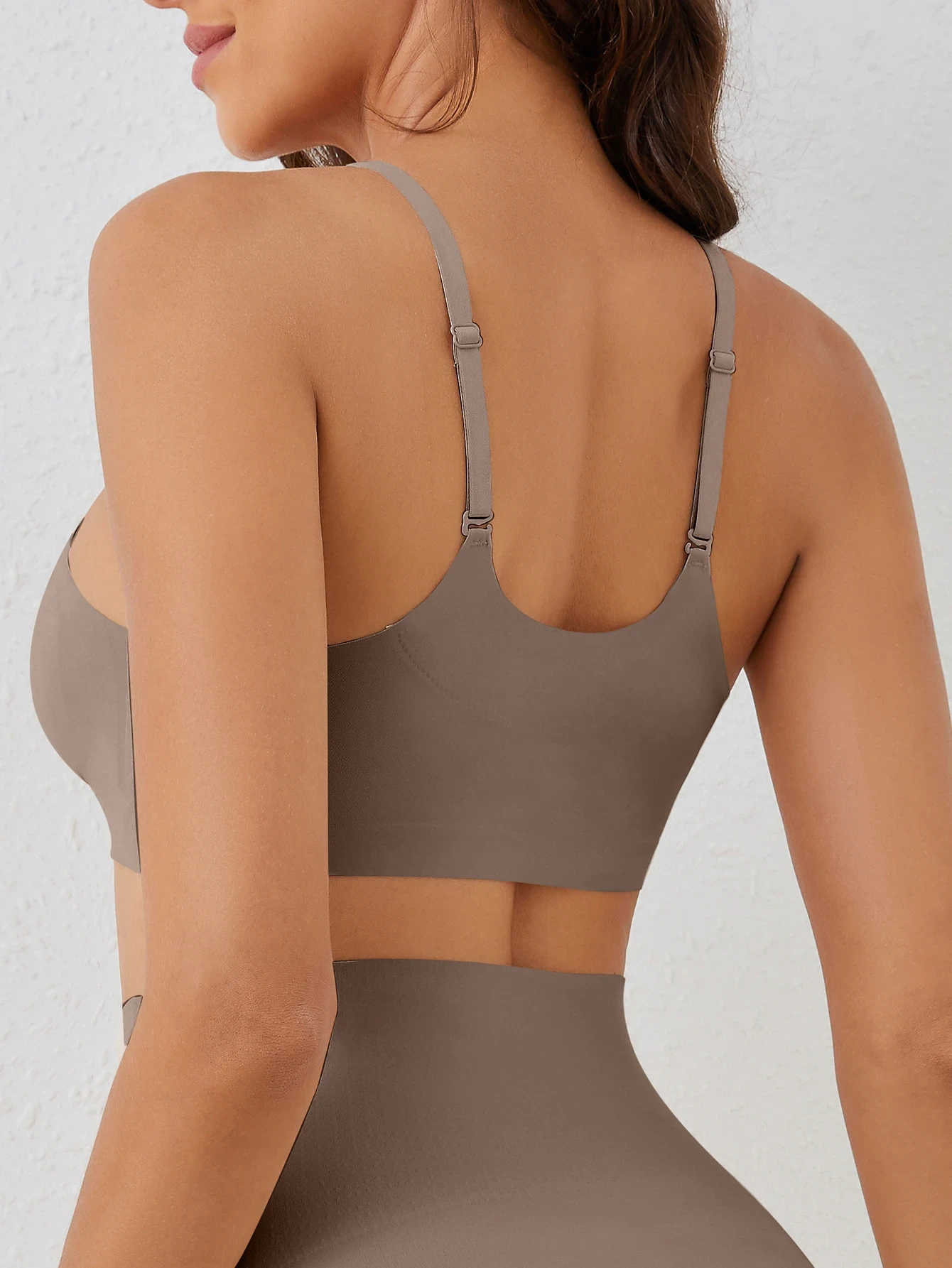 A woman shows the back of a taupe-colored bra with adjustable straps, against a white textured wall backdrop.