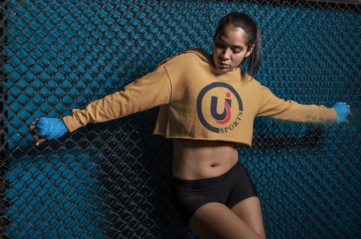 Athletic figure in a cropped sweatshirt and shorts, posing against a blue background with a black fence.