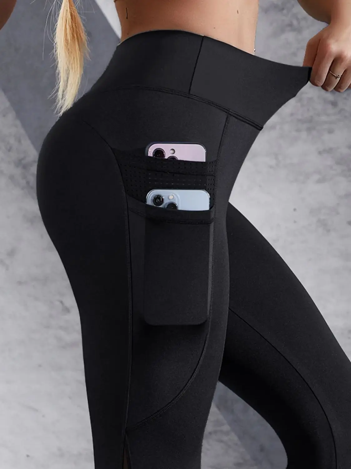 A close-up of a woman wearing black leggings featuring side pockets holding two smartphones, showcasing style and functionality.